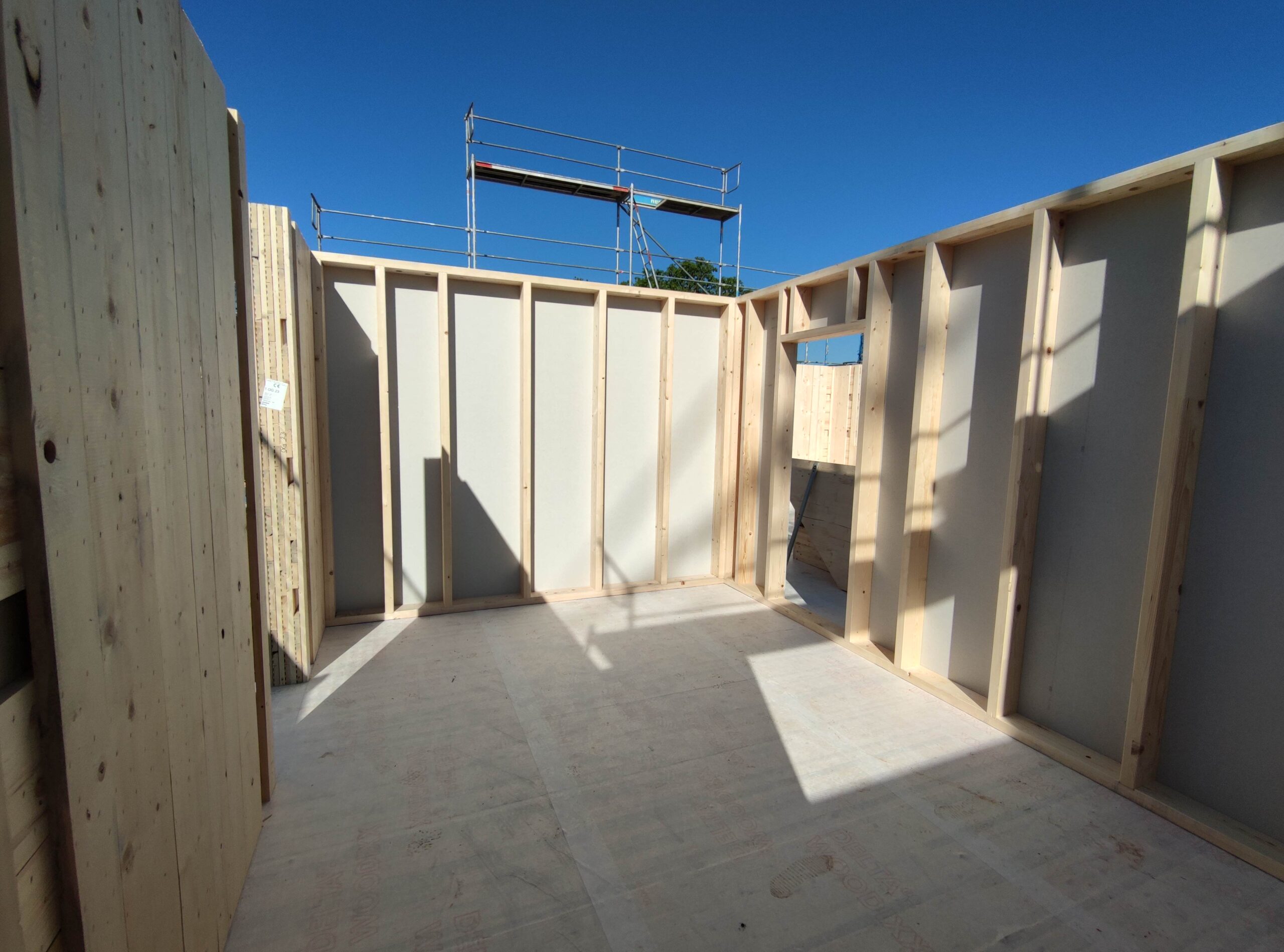 Interior view of a room under construction with exposed wooden studs and framing, doorway opening, and blue sky visible outside.